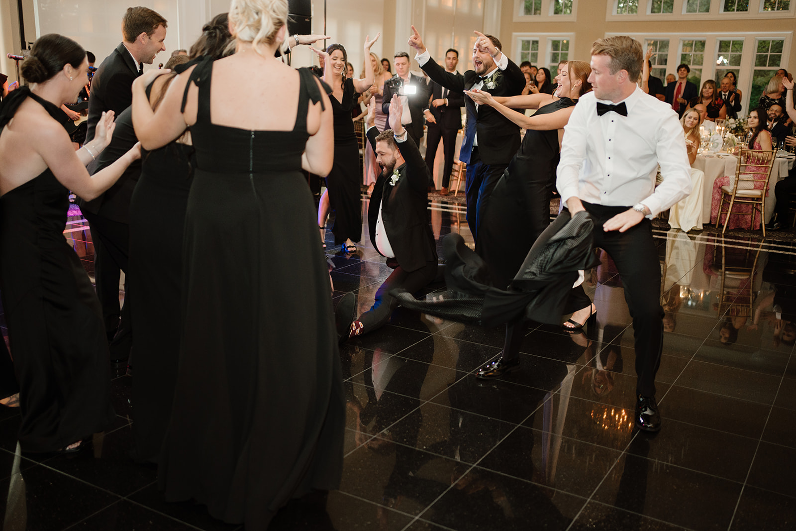 Groomsman doing a full split on the glossy reception dance floor as surrounding wedding guests cheer and laugh.